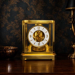 May include: A gold-toned mantel clock with a rectangular glass case and a white circular dial featuring gold-colored hour markers and hands. The clock displays the numbers 12, 3, 6, and 9, with a visible internal mechanical movement mechanism.