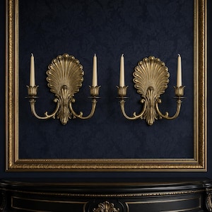 May include: Two gold-toned metal wall sconces, each featuring a central shell-shaped backplate and two curved arms holding ivory taper candles. The sconces are mounted symmetrically on a dark blue wall within a large, ornate gold-toned frame.