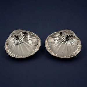 May include: Two silver-toned dishes shaped like open scallop shells. Each dish features a reflective, metallic surface with deep, radiating fluted grooves that extend from the central hinge area to the scalloped, curved outer edges.