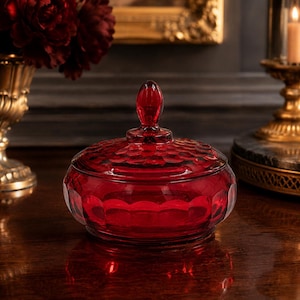May include: A round, deep red glass candy dish with a matching lid. The dish features a textured, scalloped pattern along the sides and lid, topped with a smooth, oval-shaped finial handle. The glass is translucent and reflects light.