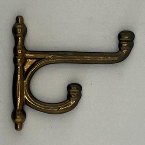May include: A vintage-style brass wall hook with a decorative design. The hook features a curved lower section and two rounded ends, with a vertical support. The brass has a warm, aged appearance, suggesting it is an antique or vintage piece.