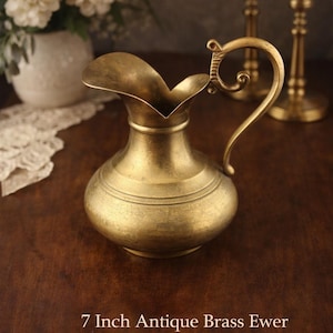May include: Antique brass ewer, approximately 7 inches tall, with a distinctive spout and ornate handle. The jug has a rounded body with horizontal bands and is displayed on a dark wooden surface. The text "7 Inch Antique Brass Ewer" is at the bottom.