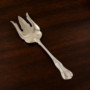 May include: A silver-toned serving fork featuring a wide, three-pronged head with a curved, flame-like design. The handle is decorated with embossed shell motifs and intricate scrollwork patterns, extending into a slender, polished neck.