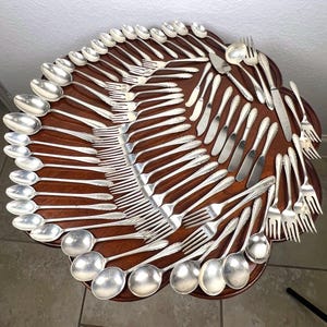 May include: A collection of silver-colored flatware, including spoons, forks, and knives, arranged on a dark wooden table. The silverware is neatly organized, showcasing a classic dining set.