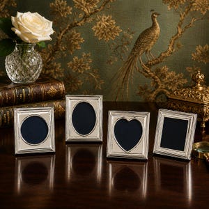 May include: A set of four silver-toned metal picture frames with varying aperture shapes including a circle, an oval, a heart, and a square. Each frame features a stepped border design and stands upright on a dark surface, displaying dark blue inserts.