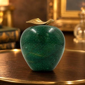 May include: A polished dark green stone apple figurine with a smooth, speckled texture and subtle natural veining. The decorative fruit features a metallic gold-toned leaf attached to the top, creating a contrast between the stone and metal.