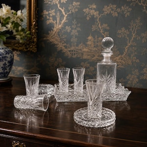 May include: A crystal bar set with a decanter, shot glasses, and serving trays. The glassware features a starburst pattern. The set is displayed on a dark wooden surface, with a floral vase and mirror in the background.