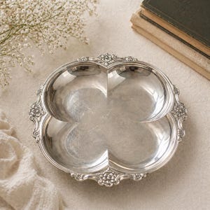 May include: Silver-colored, four-section serving dish with a clover-like design. The dish has a decorative, floral-patterned edge. The dish is on a neutral-colored surface, with a stack of books and flowers in the background.