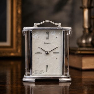 May include: A rectangular Bulova mantel clock with a silver-plated frame and a handle on top. The clock face features a white pearlescent finish, black Roman numerals at the twelve and six o'clock positions, and the text Bulova and Silverplate.