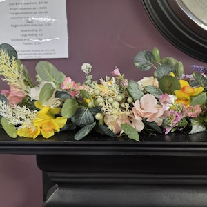 May include: A decorative floral garland featuring yellow daffodils, pink roses, and green leaves. The garland is displayed on a black surface, showcasing a mix of artificial flowers. The arrangement includes a variety of textures and colours.