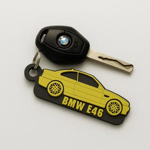 Może przedstawiać: Czarny kluczyk do samochodu BMW oraz żółto-czarny brelok do kluczy w kształcie samochodu BMW E46. Brelok posiada żółty wzór samochodu z czarnymi kołami i napisem "BMW E46". Kluczyk posiada logo BMW.