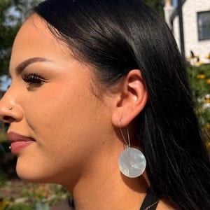 Pode incluir: Um par de brincos circulares pendentes de cor branca iridescente com um gancho de fio prateado. Os brincos apresentam uma superfície lisa e refletora que capta a luz, suspensos por um simples laço de metal que passa pelo lóbulo.