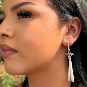 May include: A silver-toned earring featuring a small hummingbird charm suspended from a hook, with two long, slender, white cylindrical pendants hanging vertically from the bird, creating a delicate and elongated drop design.