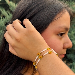 May include: A double-strand bracelet with white beads and small yellow flower accents. The bracelet is worn on a wrist, with the background showing green foliage and a person's arm and face.