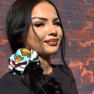 Puede incluir: Una colorida scrunchie con un diseño de dibujos animados que presenta una variedad de colores, incluyendo azul, amarillo y rojo. La scrunchie está envuelta alrededor de una cola de caballo. El fondo es una pared de ladrillos con un mural.