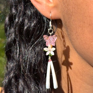 May include: A dangling earring featuring a pink butterfly, a white flower, and two white, elongated beads. The earring is silver-toned and hangs from a hook. The background is a blurred green.