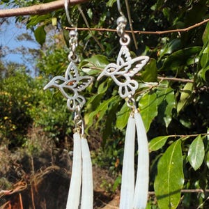 May include: Silver-toned hummingbird earrings with dangling white accents. The earrings feature a detailed hummingbird design and are suspended from a silver hook. The white accents are long and flowing, adding movement to the jewelry. The earrings are displayed against a natural background.