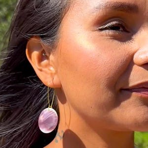 以下が含まれることがあります： 女性が身につけているのは、楕円形のピンク色のイヤリングです。イヤリングは細い金色のワイヤーで吊り下げられています。イヤリングはピンクと白のマーブル模様です。背景は、木々と水面のあるぼやけた屋外の風景です。