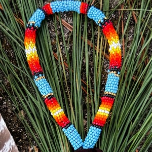 May include: A beaded lanyard with a vibrant pattern of blue, red, orange, and yellow beads. The lanyard has a black cord, a brown leather piece, and a gold-colored ring. The background includes a feather and green foliage.
