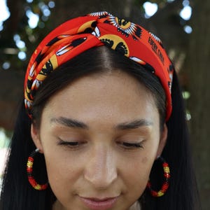 Puede incluir: Una diadema roja con un patrón repetido de diseños negros y amarillos y el texto "EVERY CHILD". La diadema se usa con pendientes de aro con cuentas rojas, amarillas y negras. La imagen está tomada al aire libre.