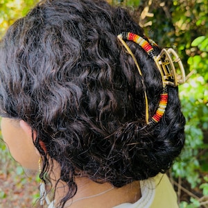 Pode incluir: Uma presilha de cabelo dourada com um detalhe de contas vermelhas, laranja, amarelas e pretas. A presilha prende cabelo escuro e encaracolado, com um fundo verde natural.