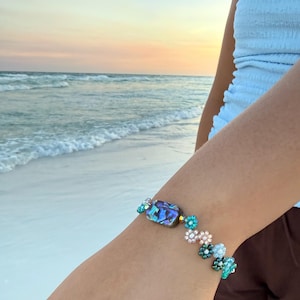 May include: A beaded bracelet featuring a central rectangular iridescent abalone shell bead flanked by small flower-shaped beads in shades of light blue, teal, and soft pink. The bracelet is constructed with small round glass beads and clear spacers.