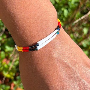 Puede incluir: Una pulsera de cuentas con cuentas blancas, negras, amarillas y rojas se lleva en la muñeca. Un anillo turquesa con un engaste plateado está en la mano. El fondo es follaje verde borroso.