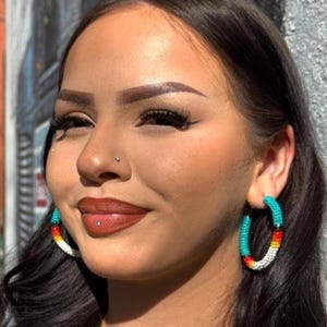 Puede incluir: Pendientes de aro con cuentas turquesas, rojas, amarillas y blancas. Los pendientes los lleva una persona con pelo oscuro y una camiseta negra. Los pendientes tienen un diseño colorido del suroeste.