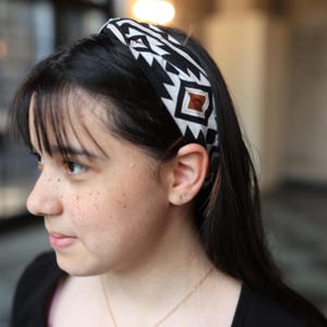 Puede incluir: Una diadema con estampado en blanco y negro con un diseño geométrico. La diadema presenta un patrón repetitivo de triángulos y cuadrados blancos sobre un fondo negro, con un cuadrado marrón en el centro. El accesorio se lleva en la cabeza.