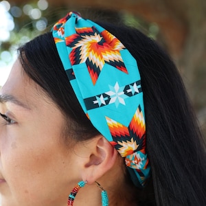 Peut inclure: Un bandeau en tissu turquoise orné d'un motif géométrique en forme d'éclat d'étoile orange, jaune et noir. Le bandeau est noué sur le dessus. Le motif se répète sur le tissu, créant un accessoire saisissant.