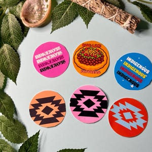 May include: A selection of circular stickers with diverse designs and text. Some stickers display the word "INDIGENOUS" in pink, blue, and white. Other stickers feature a frybread design and geometric patterns in orange, black, pink, and blue.