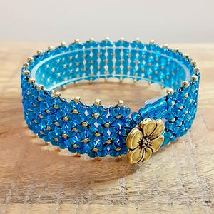 May include: A beaded bracelet in shades of turquoise and gold. The bracelet features a floral clasp and is made with small, square beads. The bracelet is approximately 2 inches in diameter.