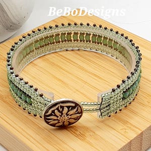 May include: A green and brown beaded bangle bracelet with a silver clasp featuring a flower design. The bracelet is on a wooden surface.