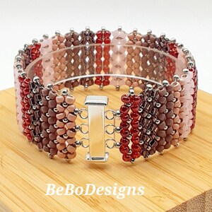 May include: A multi-colored beaded bracelet with a silver clasp. The bracelet features shades of red, pink, and brown beads arranged in a chevron pattern. The bracelet is on a wooden surface with the text "BeBoDesigns" visible.