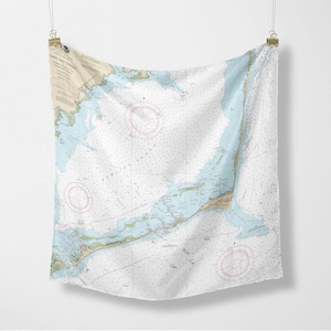 May include: A white fabric wall hanging featuring a nautical chart of Cape Hatteras. The chart includes blue, tan, and pink details, with text at the top left. The fabric is suspended by clips.