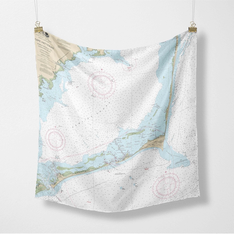 May include: A white fabric wall hanging featuring a nautical chart of Cape Hatteras. The chart includes blue, tan, and pink details, with text at the top left. The fabric is suspended by clips.