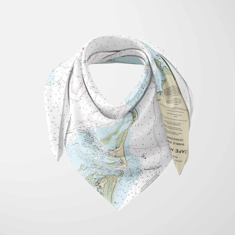 May include: A white square scarf featuring a nautical chart design with blue, green, and beige tones. The scarf is folded and draped, showcasing the detailed map of a coastal area. The scarf is made of a soft, lightweight material.