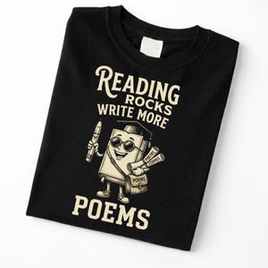 Puede incluir: Camiseta negra con un personaje de libro de dibujos animados con gafas de sol y birrete, sosteniendo un lápiz y pergaminos. La camiseta tiene el texto "Reading Rocks Write More Poems."
