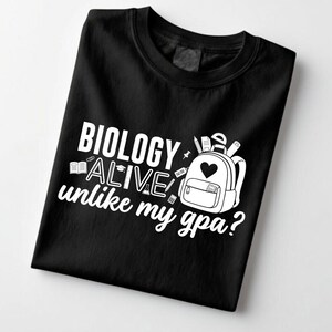 Peut inclure: T-shirt noir avec texte blanc : "BIOLOGIE ALIVE unlike my gpa ?" Un dessin de sac à dos avec un cœur, des crayons et des livres est également présent. Le t-shirt est en matière souple.