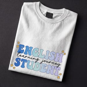 May include: White crew neck sweatshirt with the words "ENGLISH learning journey STUDENT" in blue, light blue, and purple bubble letters. Small yellow stars surround the text. The sweatshirt is folded on a black surface.