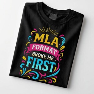 May include: Black t-shirt with the text "MLA FORMAT BROKE ME FIRST" in a colorful, decorative font. The design includes yellow, pink, blue, and teal accents. The shirt is folded neatly.