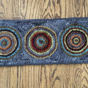 May include: A gray rug with three concentric circles in shades of brown, blue, and yellow. The circles are surrounded by a border of the same colors. The rug is on a wooden floor.