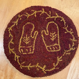 May include: Round, burgundy rug with a stitched design of two mittens and a decorative border in gold. The rug is made of a textured material and is suitable for a home decor item.