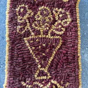 May include: A rectangular, handmade rug with a burgundy background and a gold-colored floral design. The rug has a textured surface and a gold border. The design features a vase-like shape with floral elements. The rug is likely made of wool or a similar material.
