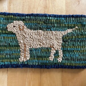 May include: A beige rug with a blue and green border features a woven image of a dog. The dog is standing on a green and blue background.