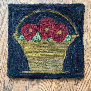 May include: A hand-hooked rug with a basket of red flowers. The basket is yellow and has a brown handle. The rug is dark blue with a black border.
