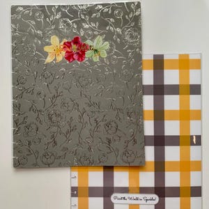 May include: Two notebooks. The top notebook has a silver floral design with colorful flower accents and the text "The Daily Mate". The bottom notebook has a yellow, brown, and white plaid pattern with the text "Paint the World in Sparkles!"