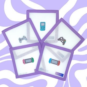 May include: Five white square needlepoint coasters with purple borders, each featuring a different video game controller design in cross-stitch. The controllers include a Game Boy, a PlayStation controller, and a Nintendo Switch.