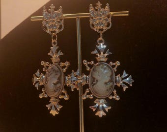 Puede incluir: Un par de pendientes de tono dorado con un diseño de camafeo. Cada pendiente presenta un camafeo de la cara de una mujer rodeado de un diseño de filigrana de tono dorado. Los pendientes cuelgan de una cadena de tono dorado con una corona en la parte superior.