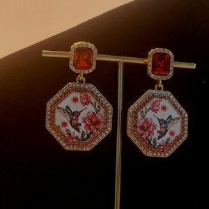 May include: A pair of gold earrings with a red gemstone and a white octagon-shaped pendant featuring a hummingbird and floral design.